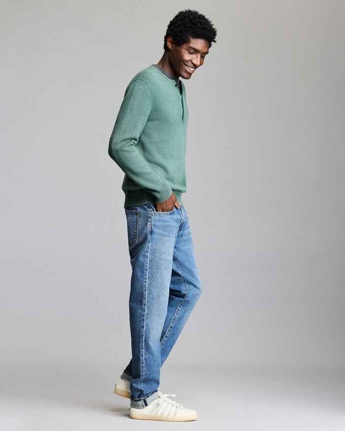 A man stands in profile smiling, wearing a green long-sleeve shirt, blue jeans, and white sneakers with stripes, against a plain gray background.