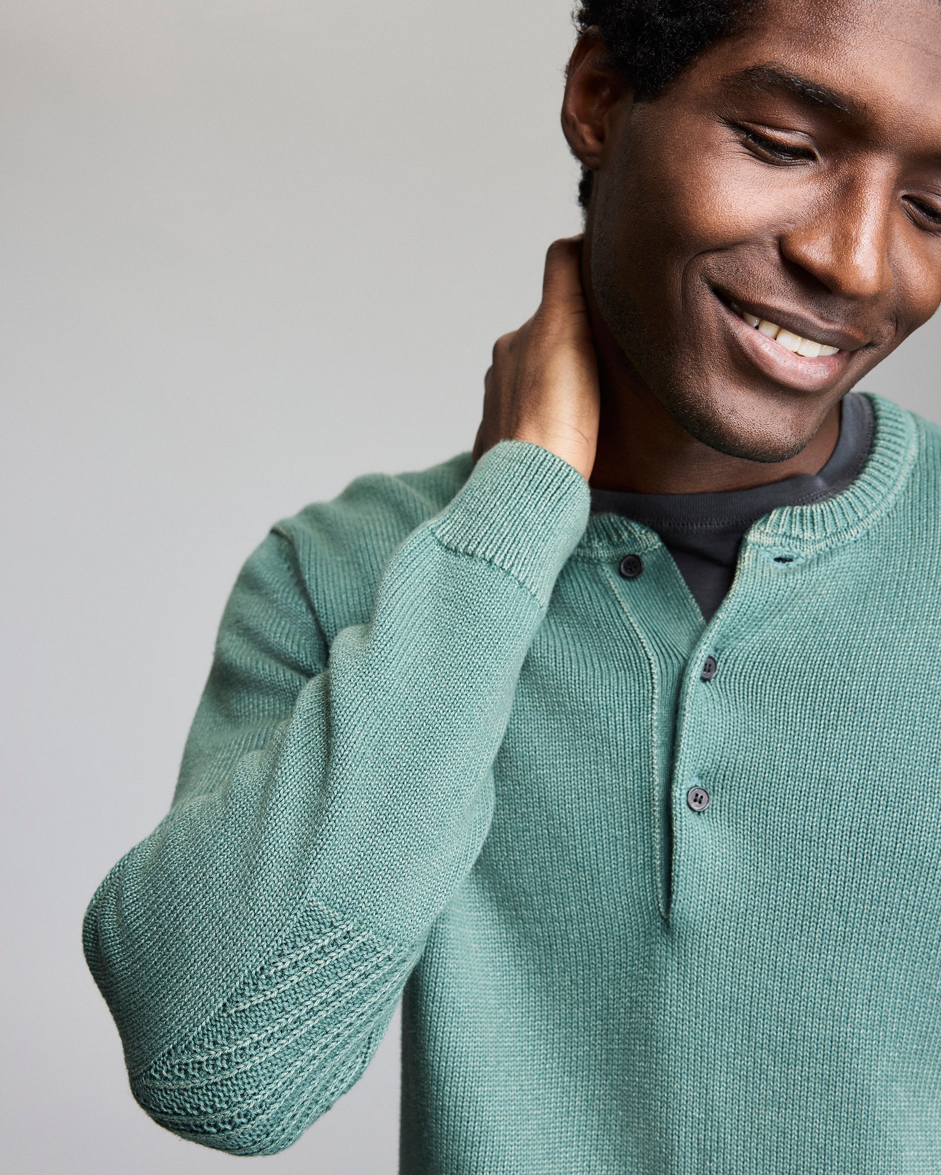 A person wearing a green knit henley sweater smiles and touches the back of their neck, looking down. The background is plain and light-colored.
