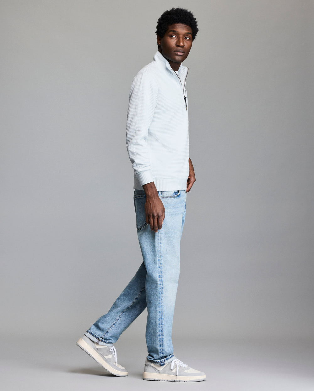 A man wearing a light-colored quarter-zip sweater, light blue jeans, and light gray sneakers stands and looks to his side against a plain, neutral background.