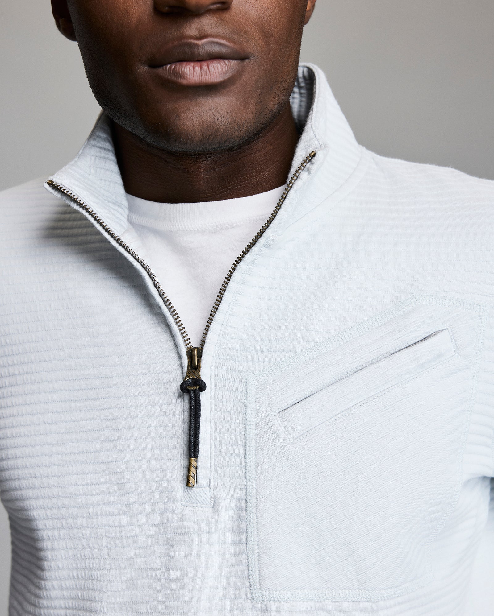 A close-up of a person wearing a white textured quarter-zip pullover over a white t-shirt. The pullover features a visible chest pocket and a partially unzipped zipper. The background is plain and neutral.