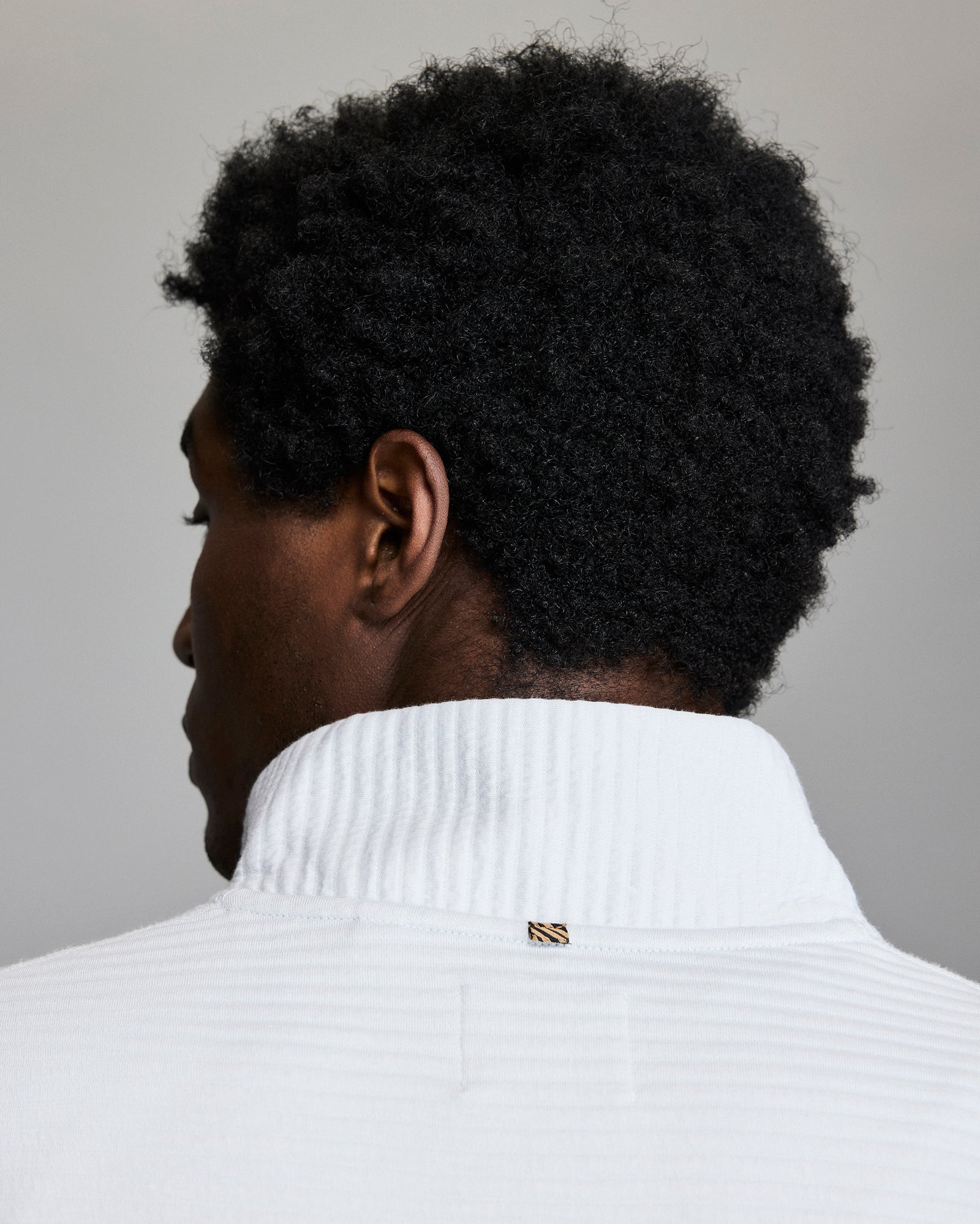 A person with short curly hair is shown from behind, wearing a white textured collared shirt with a small gold and black tag at the back. The background is plain and neutral.