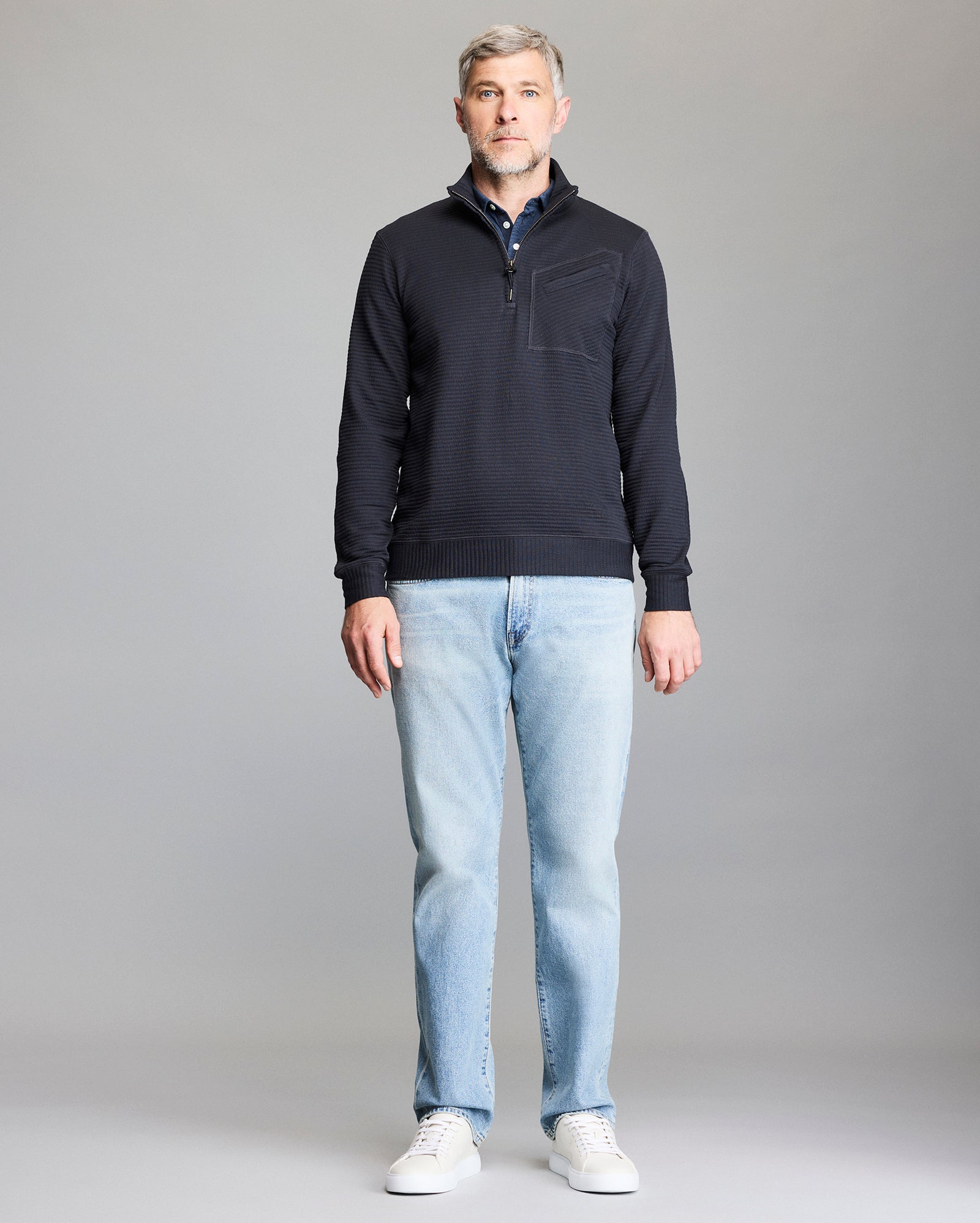 A man with short gray hair and a beard stands facing forward, wearing a navy quarter-zip pullover, light blue jeans, and white sneakers against a plain gray background.