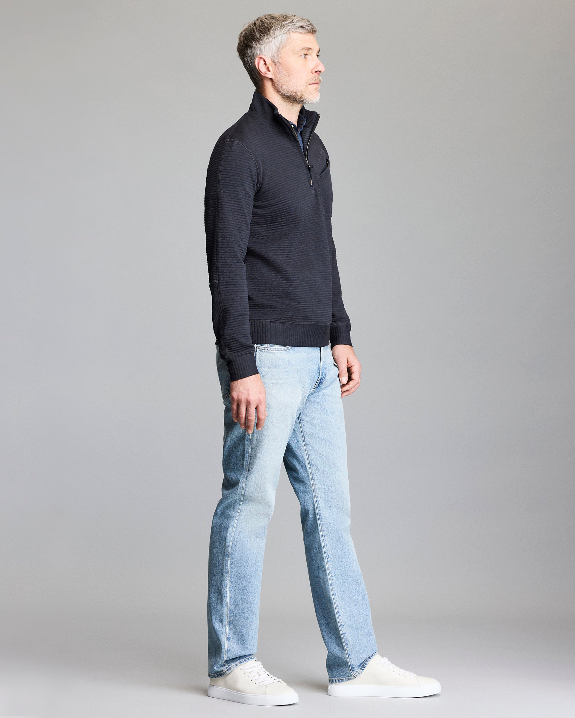 A man with gray hair stands sideways, wearing a dark zip-up sweater, light blue jeans, and white sneakers, against a plain gray background.