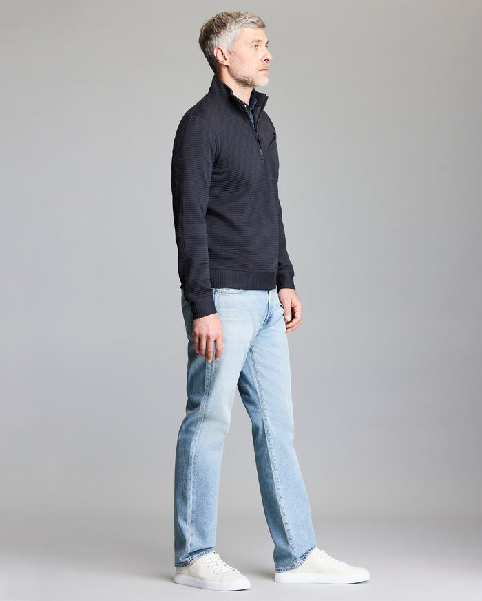 A man with gray hair stands sideways, wearing a dark zip-up sweater, light blue jeans, and white sneakers, against a plain gray background.