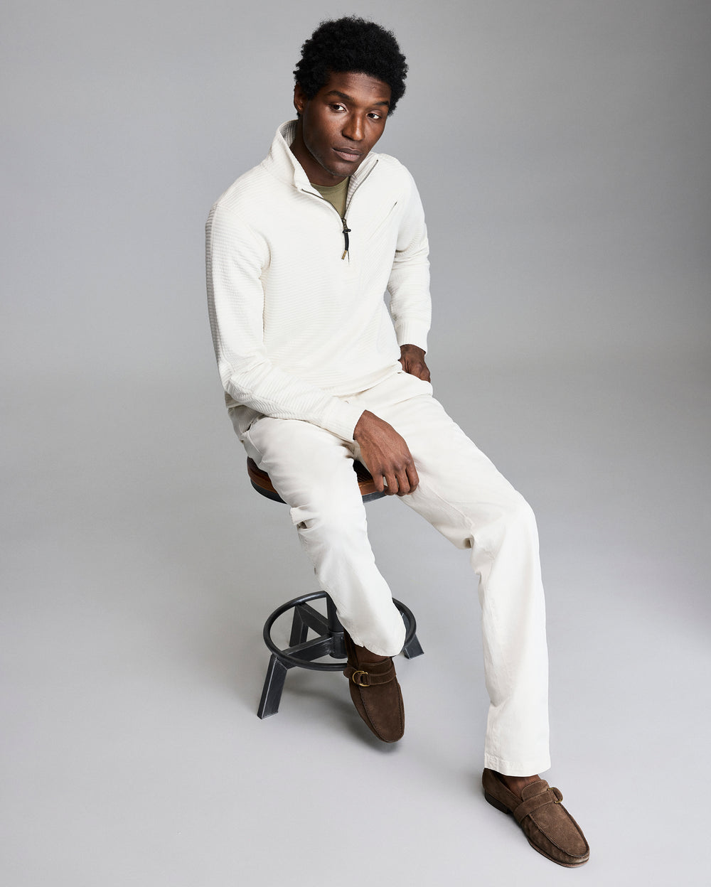 Man sitting on a stool wearing a white sweater and pants with brown shoes on a gray background
