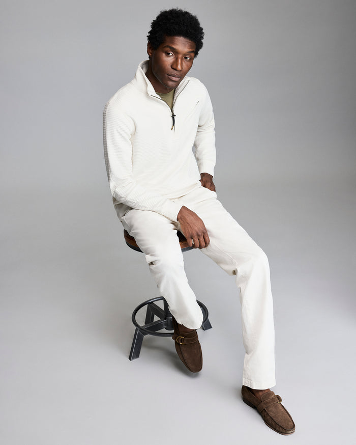 Man sitting on a stool wearing a white sweater and pants with brown shoes on a gray background
