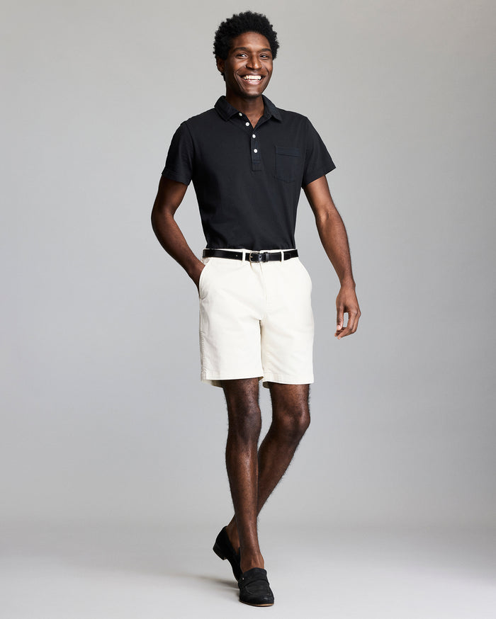 A smiling man stands against a plain gray background, wearing a black polo shirt, white shorts, a black belt, and black loafers. One hand is in his pocket and he appears relaxed and confident.