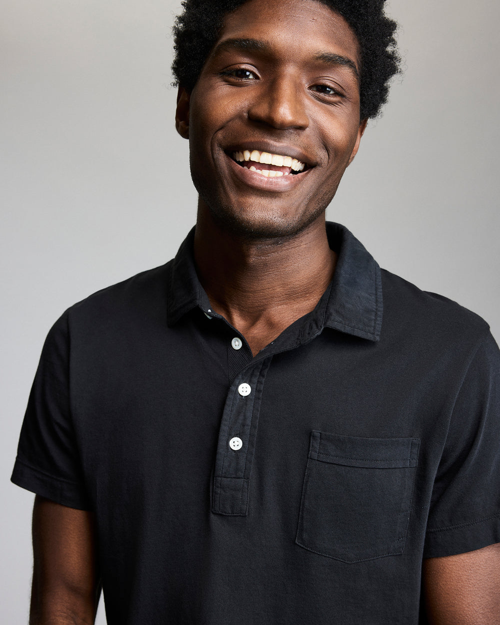 A man with short curly hair is smiling at the camera. He is wearing a black collared shirt with a front pocket against a plain, light background.
