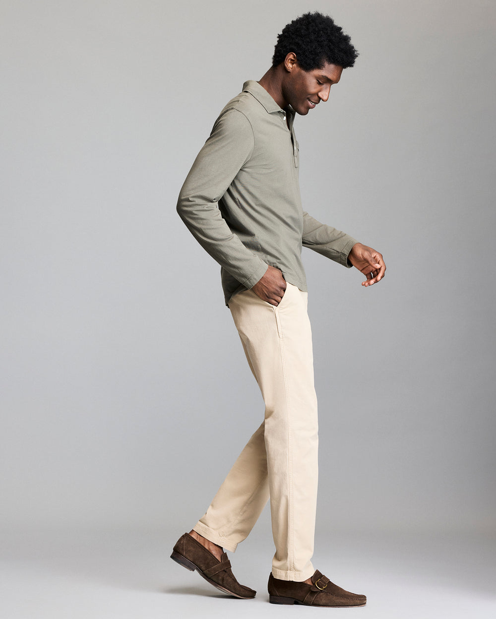 A man in a light green long-sleeve shirt, beige pants, and brown loafers walks to the right against a plain gray background, looking down with a slight smile.