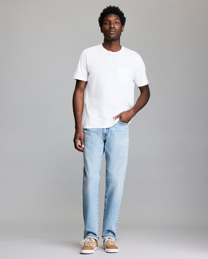 A man stands against a plain background wearing a white short-sleeve T-shirt with a chest pocket, light blue jeans, and tan sneakers. He has one hand in his pocket and a neutral expression. Alternate