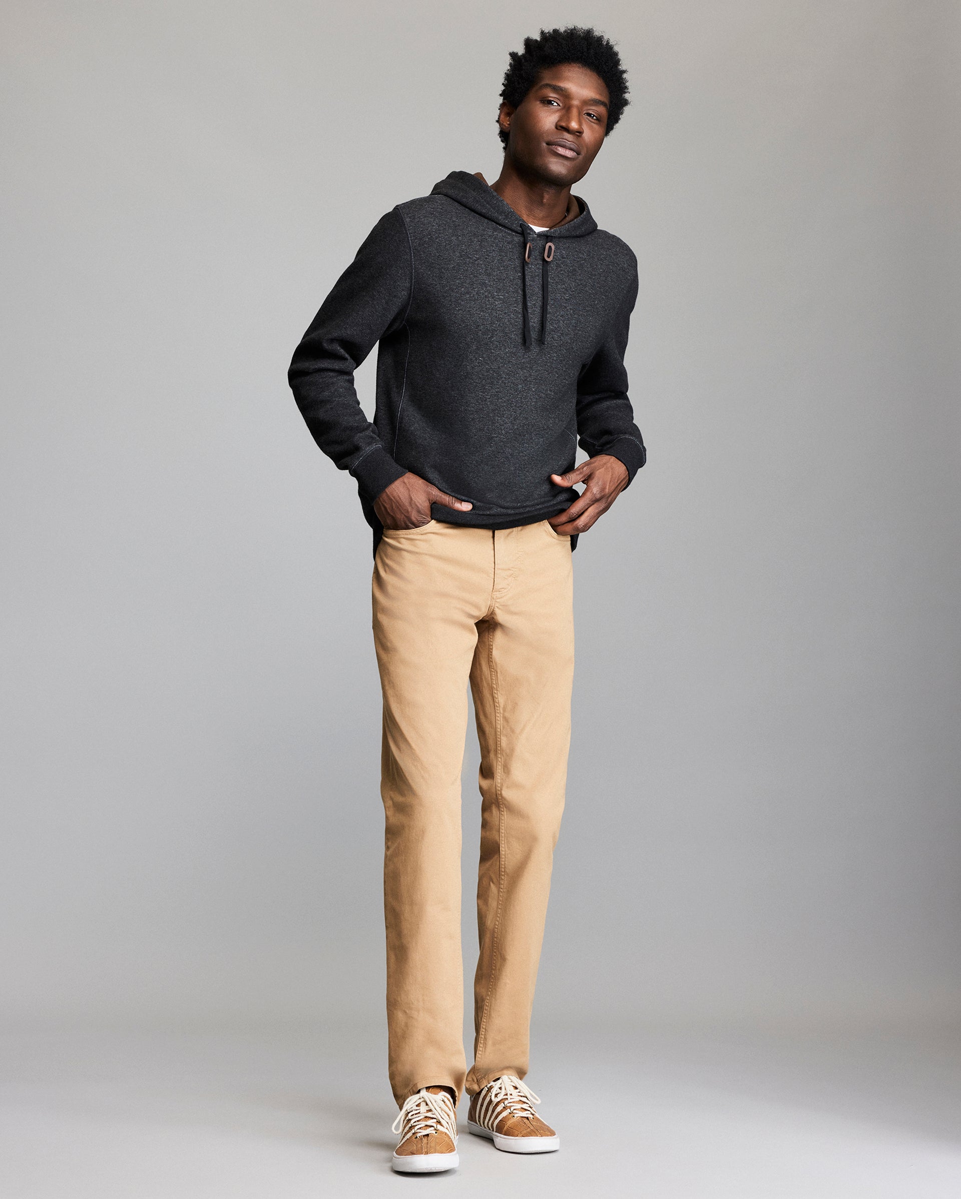 A man stands against a plain background wearing a dark gray hoodie, tan pants, and tan sneakers with white soles. He is facing forward with a relaxed posture and hands in his pockets.