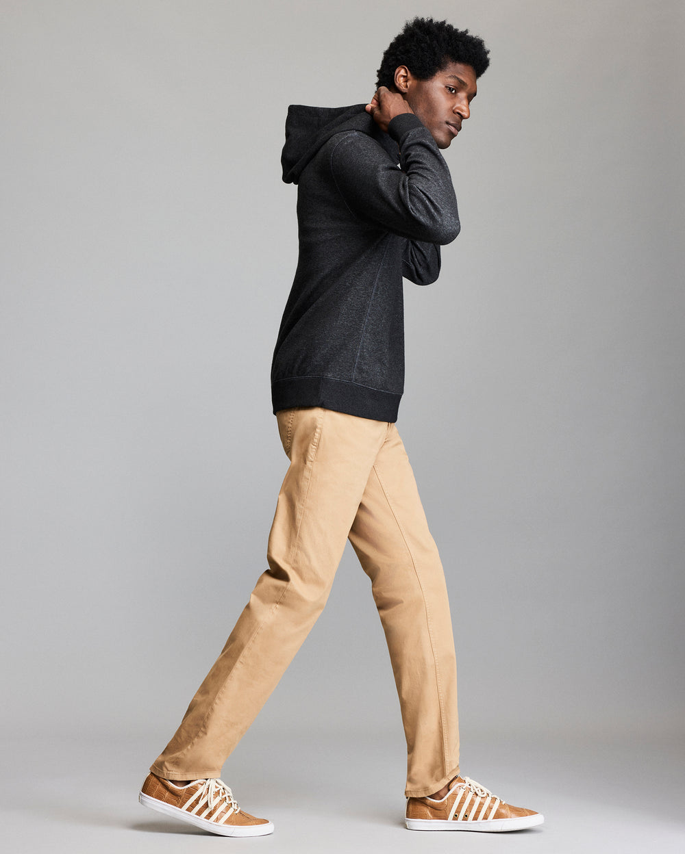 A person wearing a dark hoodie, tan khaki pants, and brown sneakers with white laces walks sideways against a plain gray background.