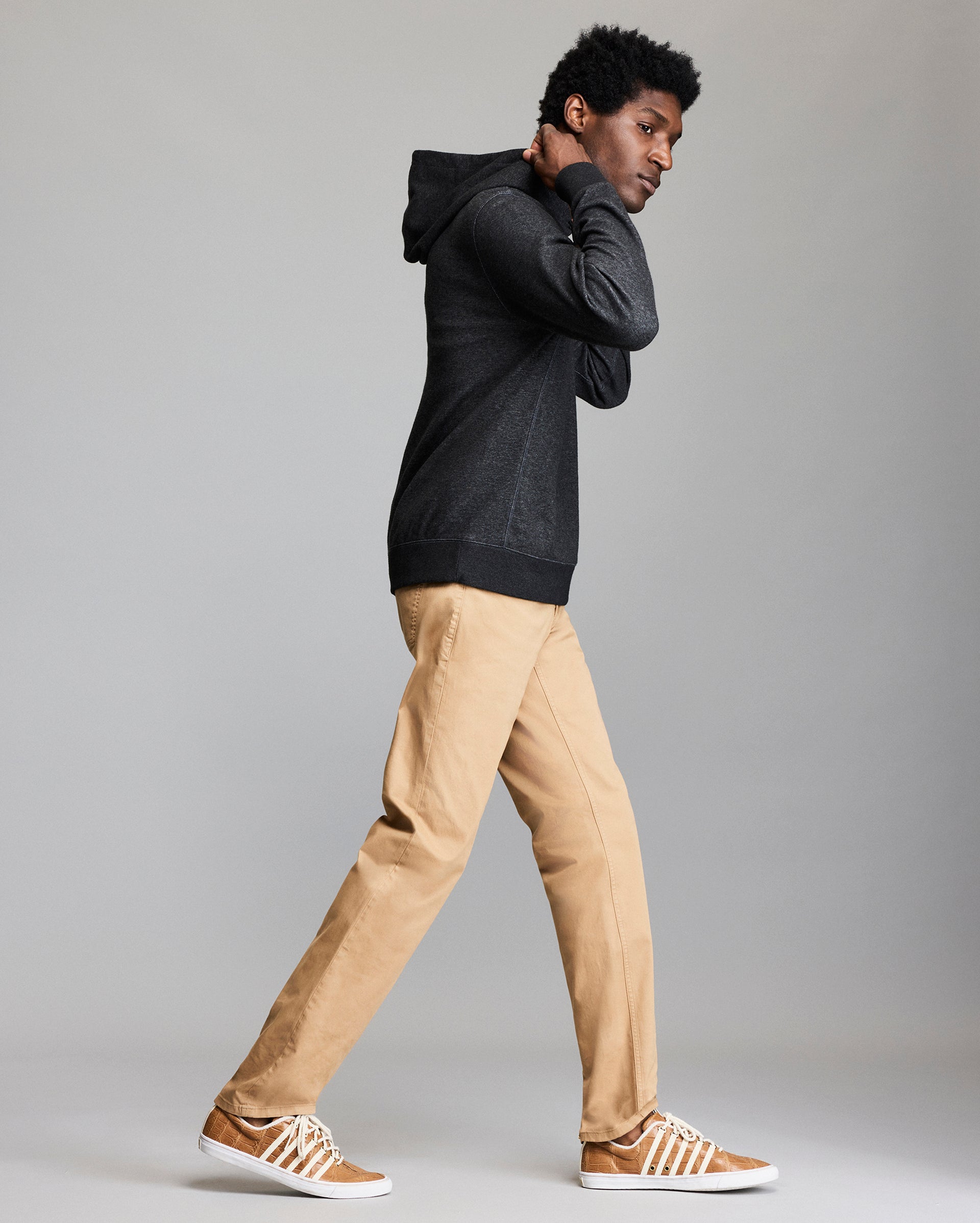 A person wearing a dark hoodie, tan khaki pants, and brown sneakers with white laces walks sideways against a plain gray background.