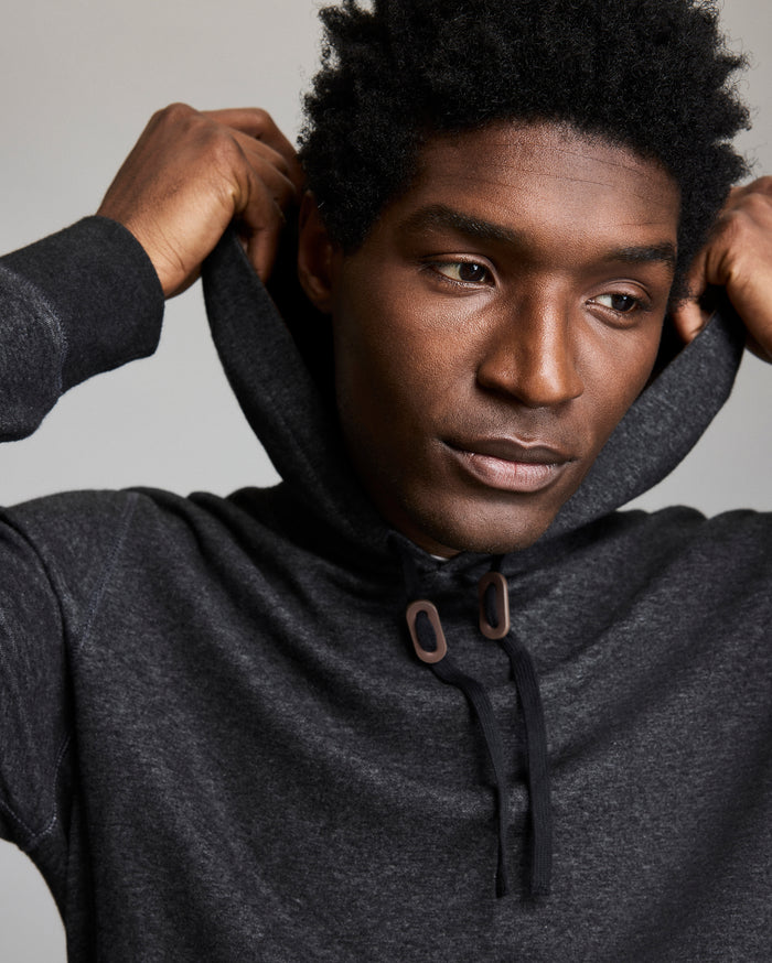 A man wearing a dark gray hoodie is pulling the hood over his head. He has short, curly black hair and a thoughtful expression, looking slightly to the side against a neutral background.