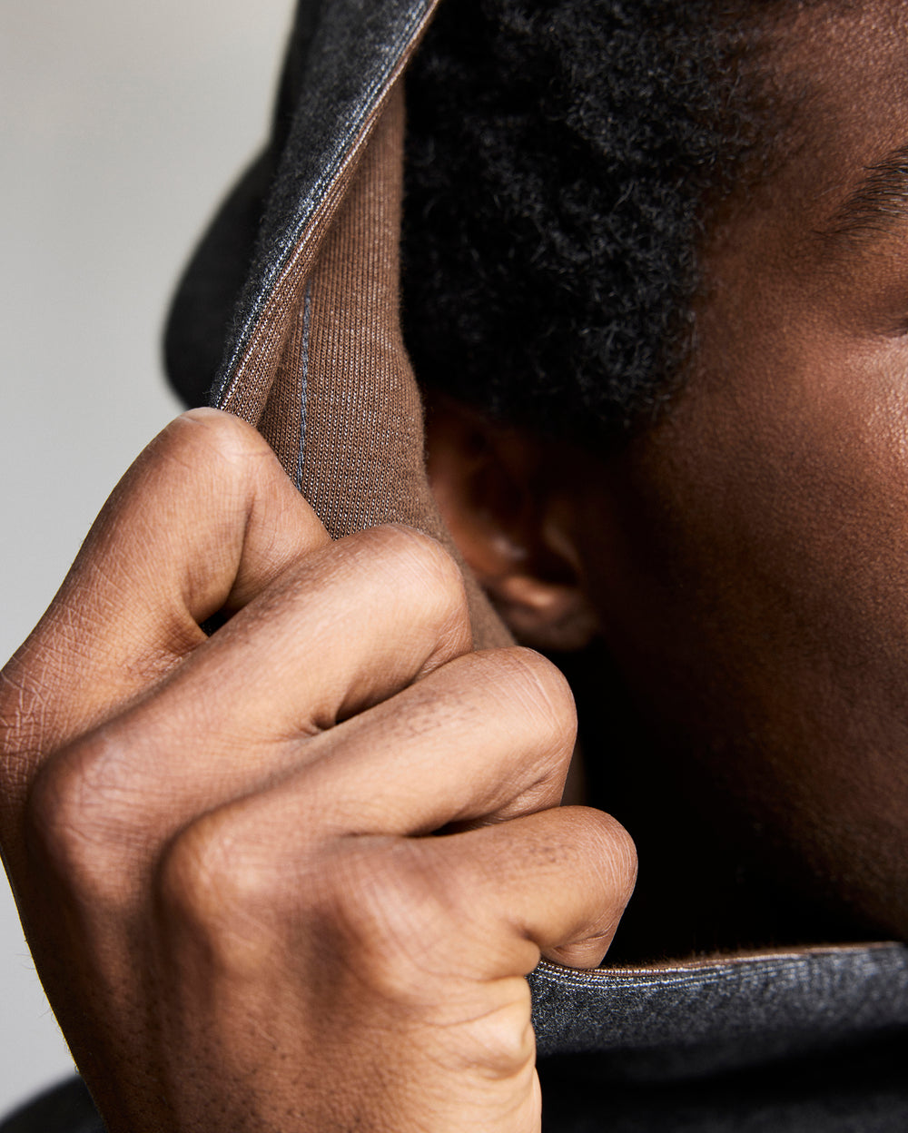 A close-up of a persons hand pulling up the hood of a grey garment, showing part of their face, ear, and textured hair. The focus is on the hand and fabric details.