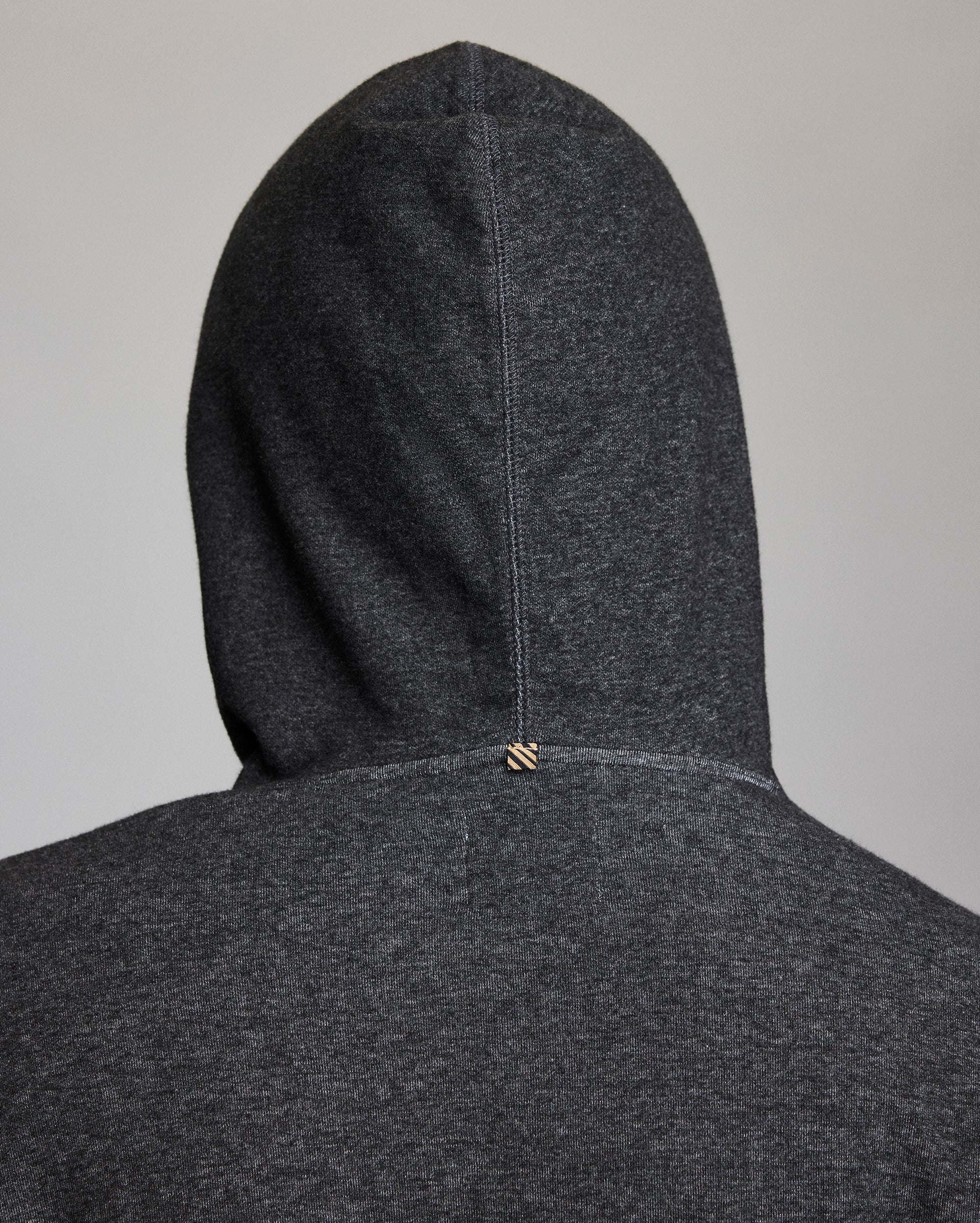 A close-up of the back of a dark gray hooded sweatshirt, showing the hood up and a small stitched tag detail at the center seam near the neckline.