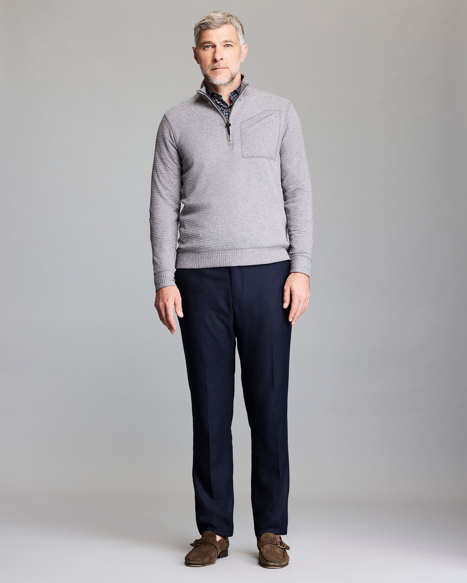A man with gray hair and a beard stands facing forward. He is wearing a light gray pullover, dark navy pants, and brown loafers against a plain gray background.
