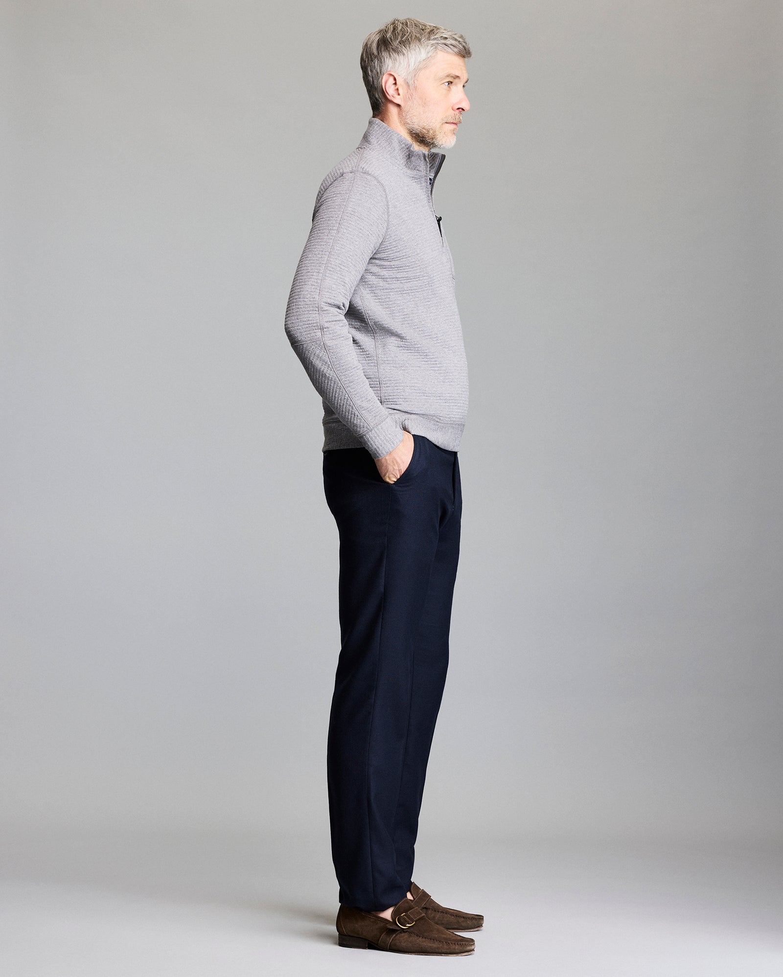 A man with short gray hair, wearing a light gray zip-up sweater, dark navy trousers, and brown loafers, stands in profile with hands in his pockets against a plain gray background.