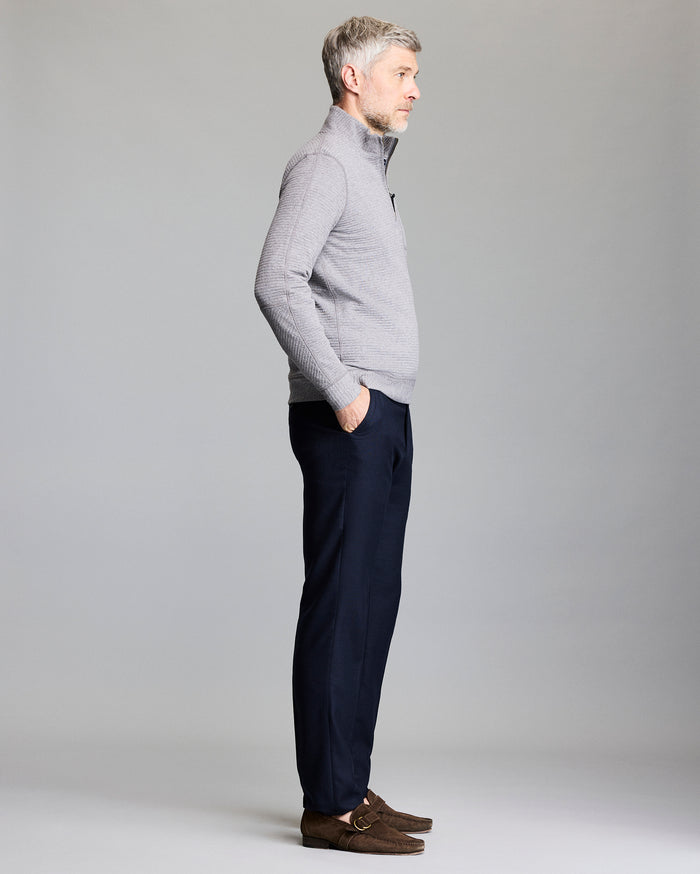 A man with short gray hair, wearing a light gray zip-up sweater, dark navy trousers, and brown loafers, stands in profile with hands in his pockets against a plain gray background.