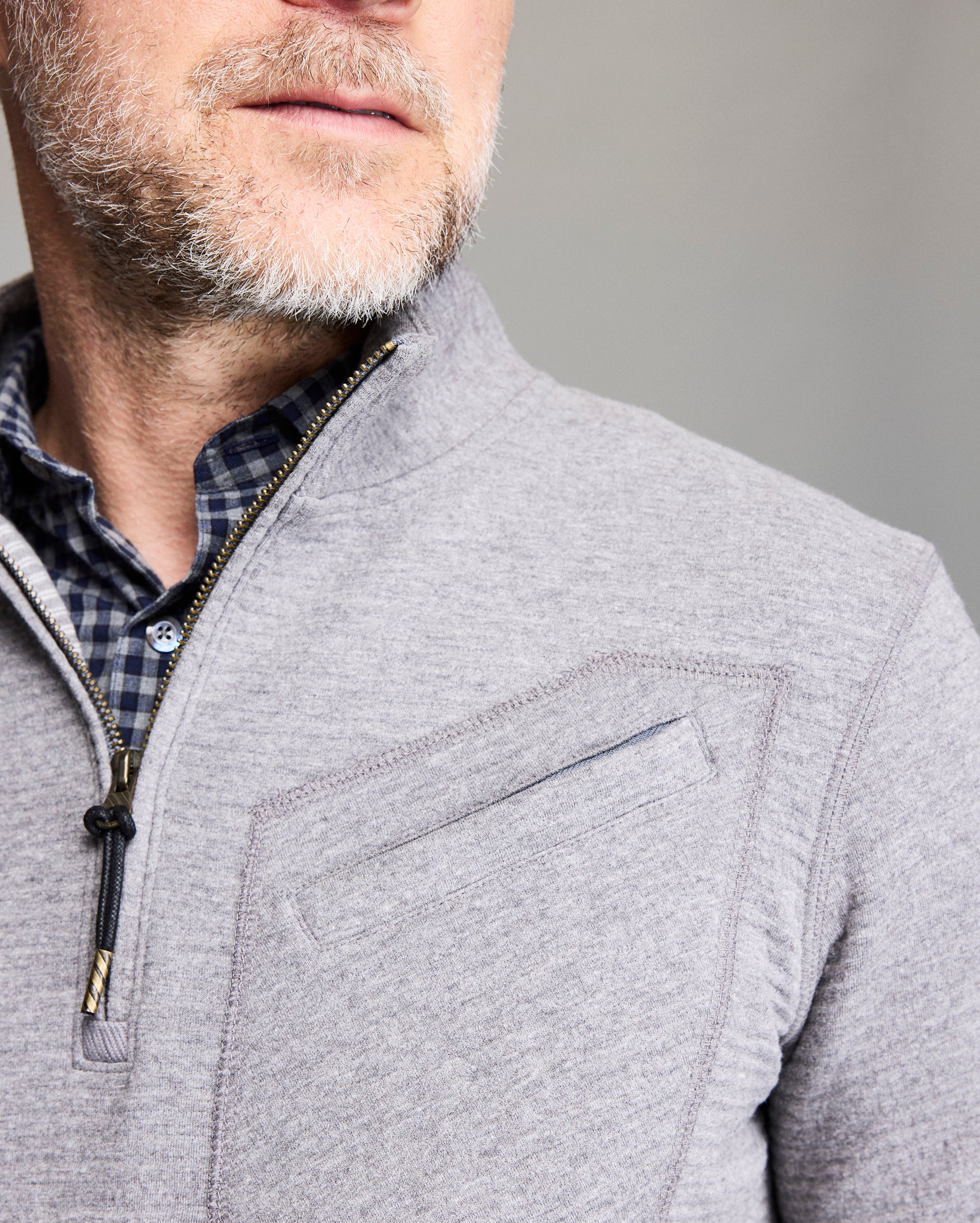 Close-up of a mans lower face and torso wearing a light gray zip-up sweater over a blue checkered shirt. He has light skin, a short gray beard, and the background is blurred.