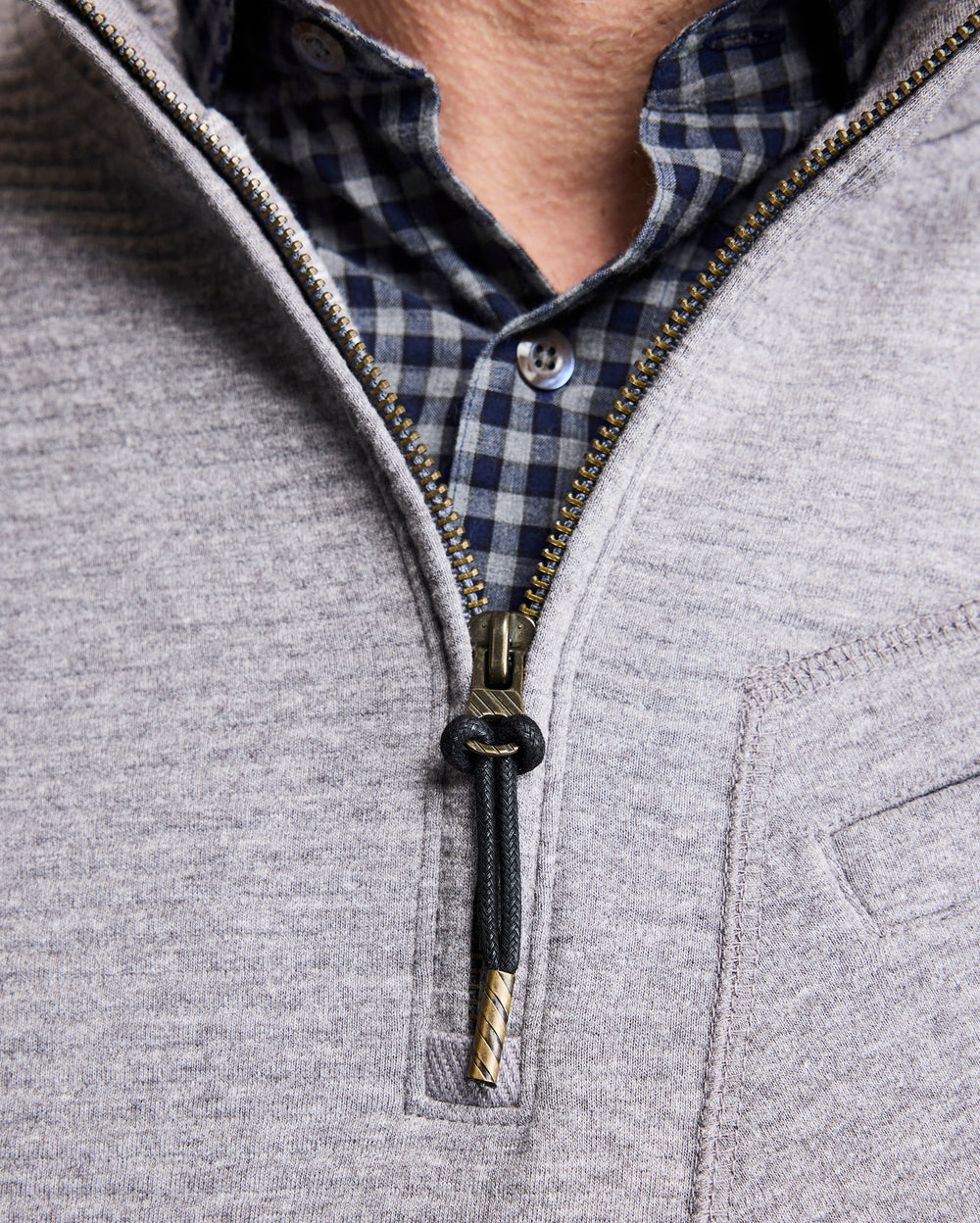 A close-up of a person wearing a blue and black checkered shirt underneath a light gray zip-up sweater, partially unzipped to reveal the shirt collar and top button.