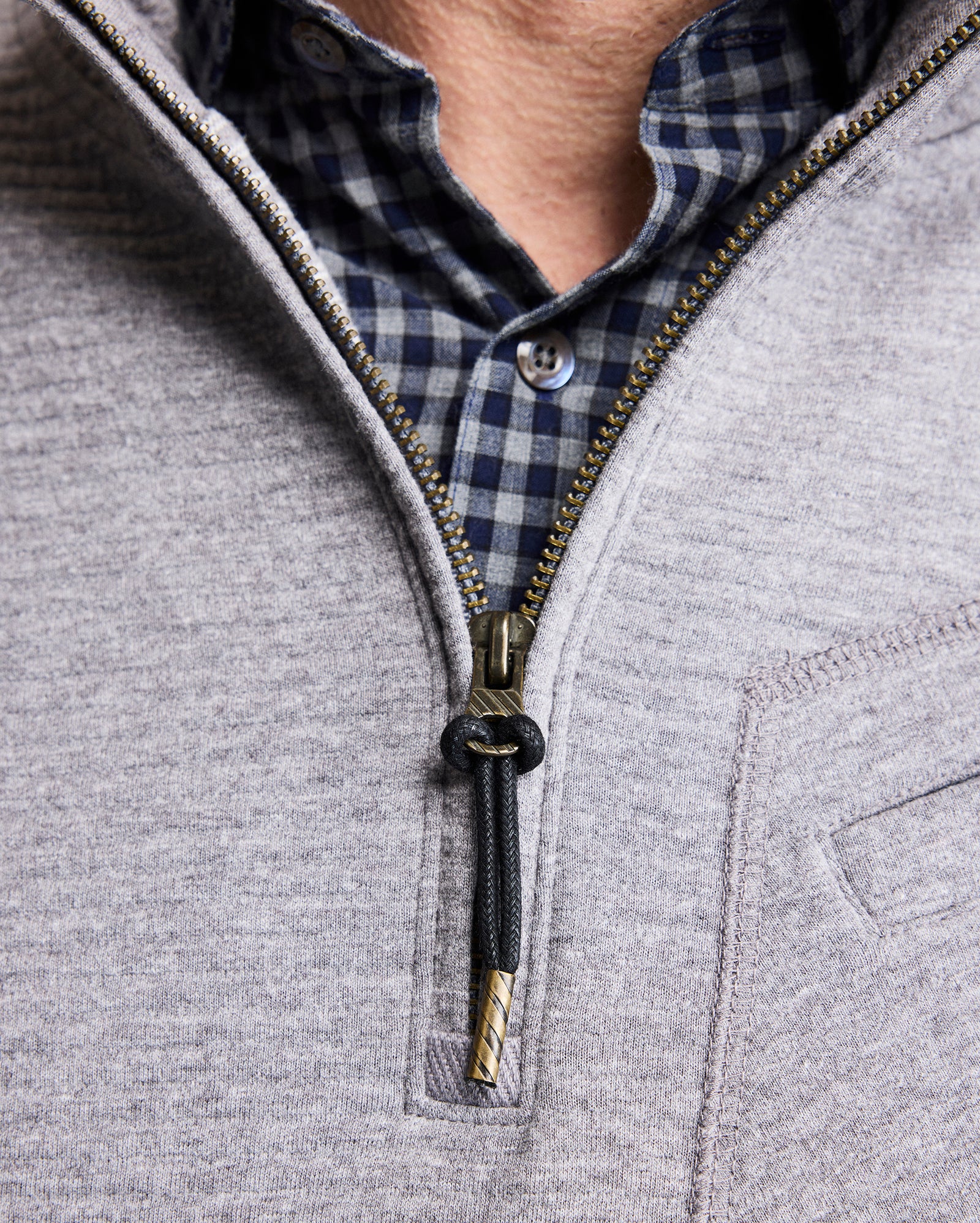 A close-up of a person wearing a blue and black checkered shirt underneath a light gray zip-up sweater, partially unzipped to reveal the shirt collar and top button.
