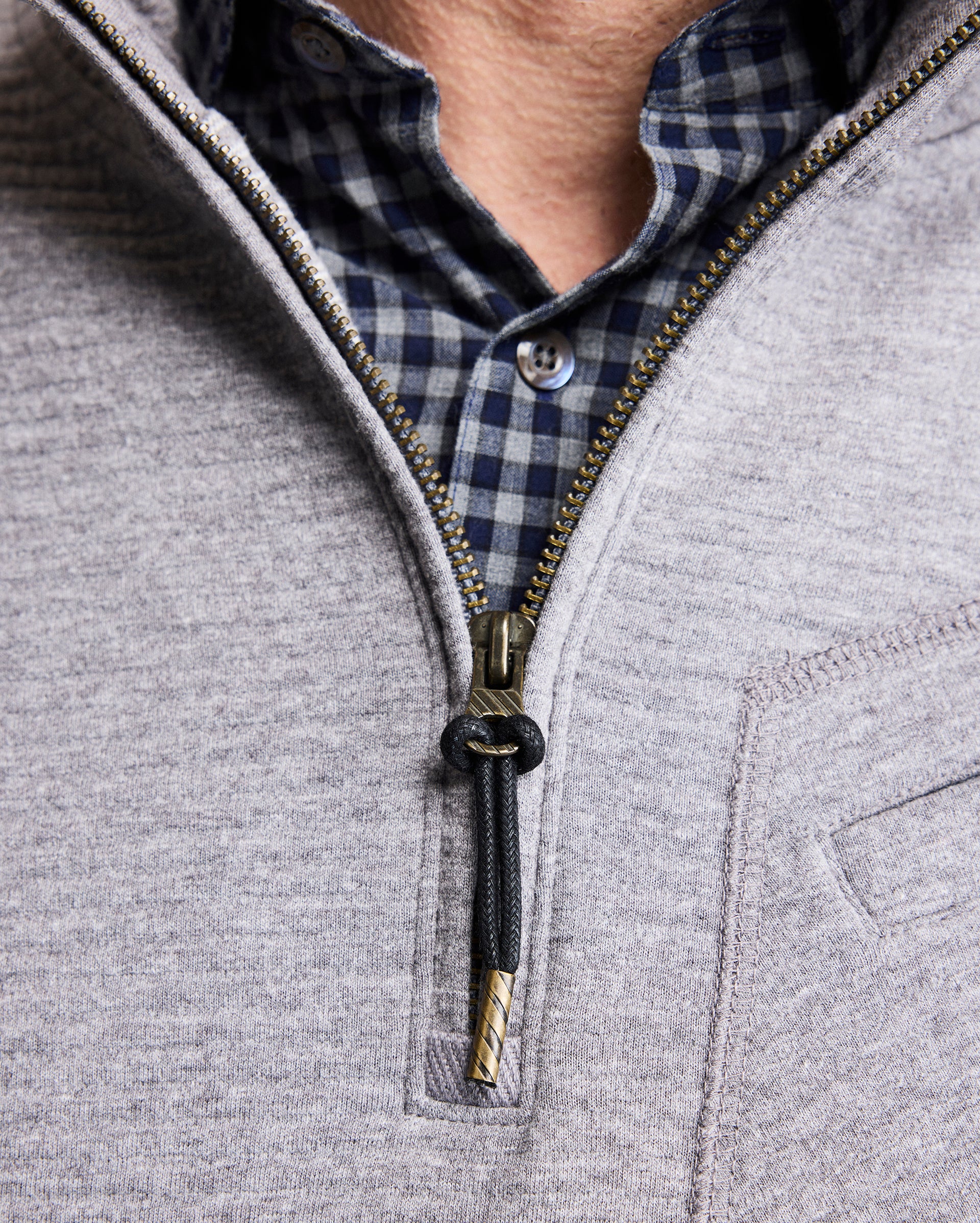 A close-up of a person wearing a blue and black checkered shirt underneath a light gray zip-up sweater, partially unzipped to reveal the shirt collar and top button.