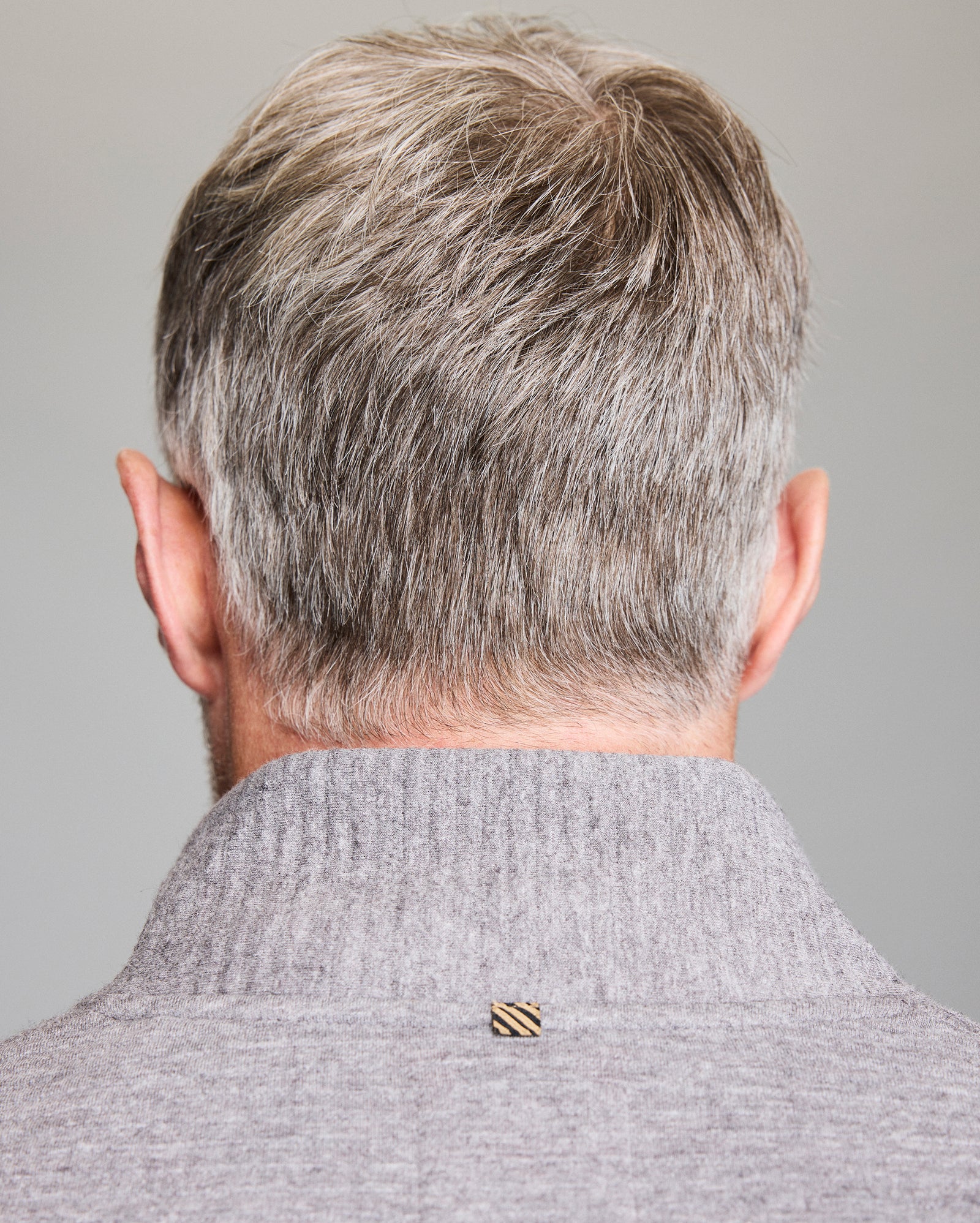 A close-up of a man with gray hair seen from behind, wearing a light gray sweater with a raised collar and a small, square, striped tag on the back.