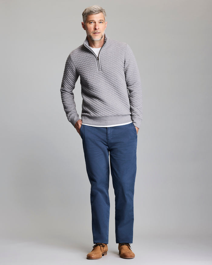 A man with gray hair, wearing a light gray quilted zip-up sweater, white shirt, blue pants, and brown shoes, stands with his hands in his pockets against a plain gray background.