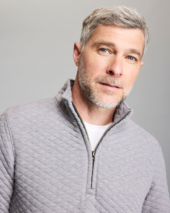 A man with short gray hair and blue eyes is wearing a light gray, quilted zip-up sweater over a white shirt. He has a short beard and is looking slightly to the right, standing in front of a plain gray background.
