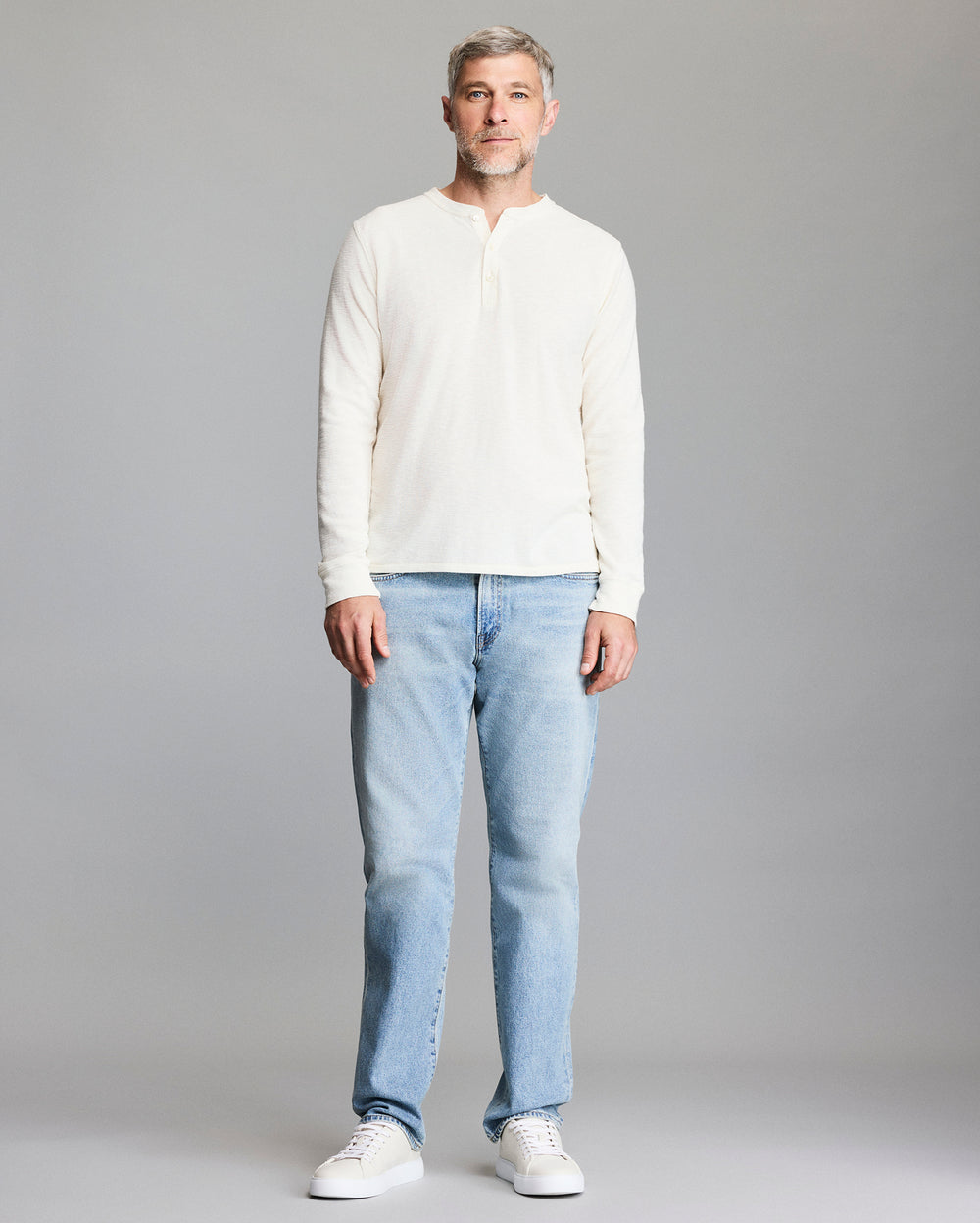 A man with short gray hair and a beard stands facing forward, wearing a cream long-sleeve henley shirt, light blue jeans, and white sneakers against a plain gray background.