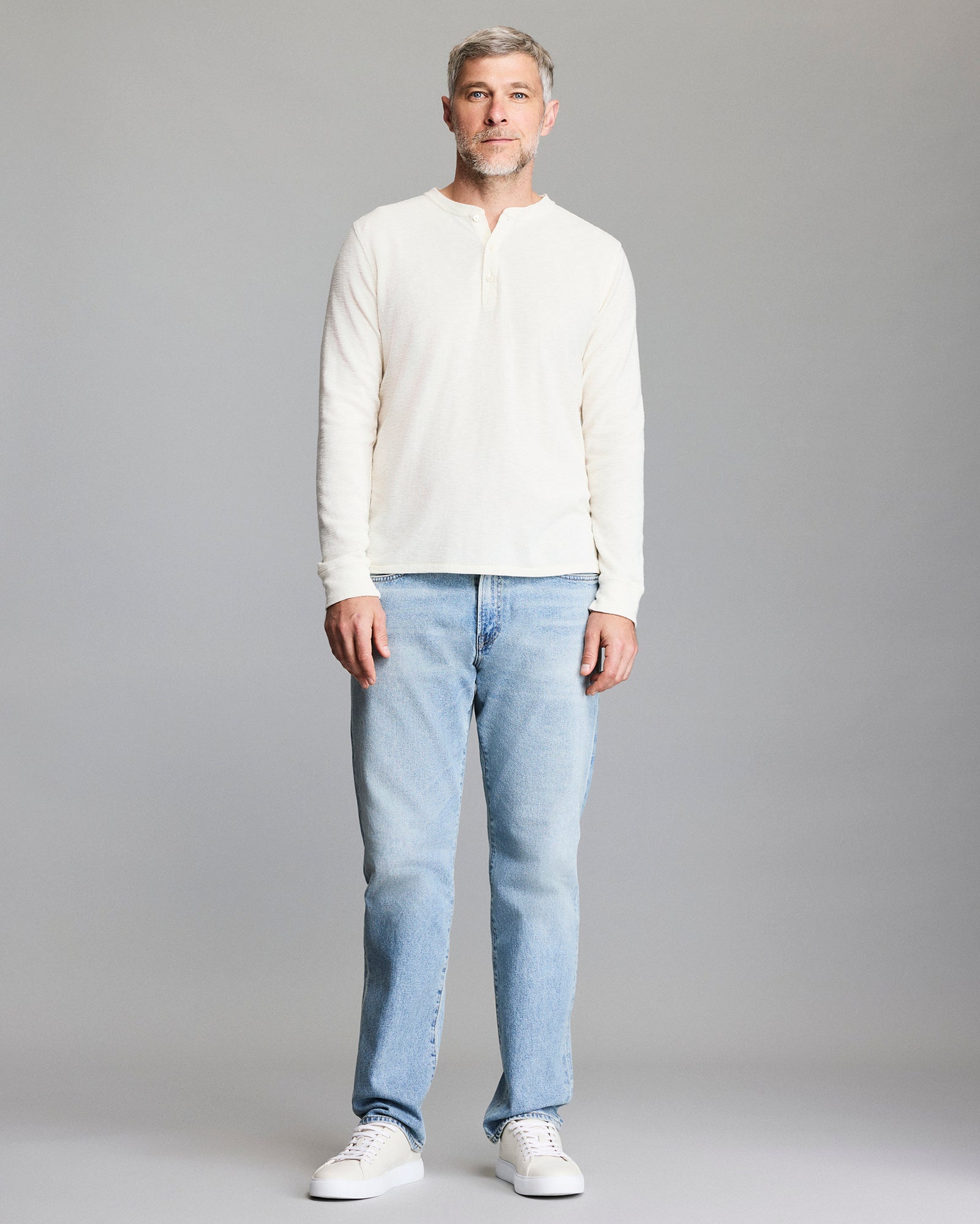 A man with short gray hair and a beard stands facing forward, wearing a cream long-sleeve henley shirt, light blue jeans, and white sneakers against a plain gray background.