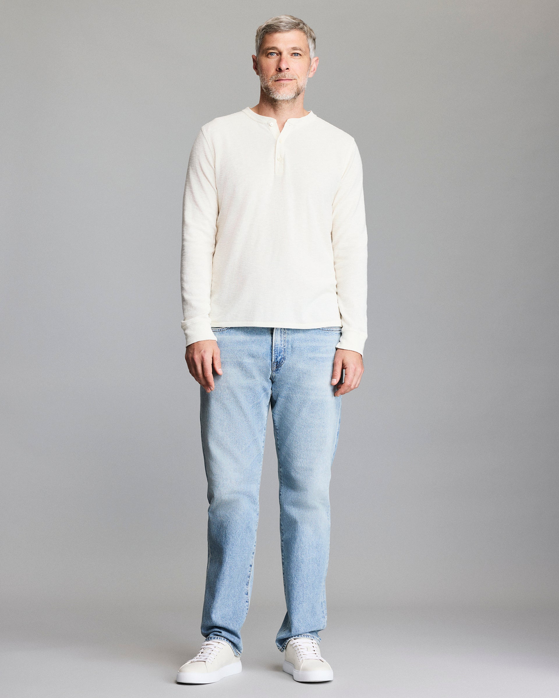 A man with short gray hair and a beard stands facing forward, wearing a cream long-sleeve henley shirt, light blue jeans, and white sneakers against a plain gray background.