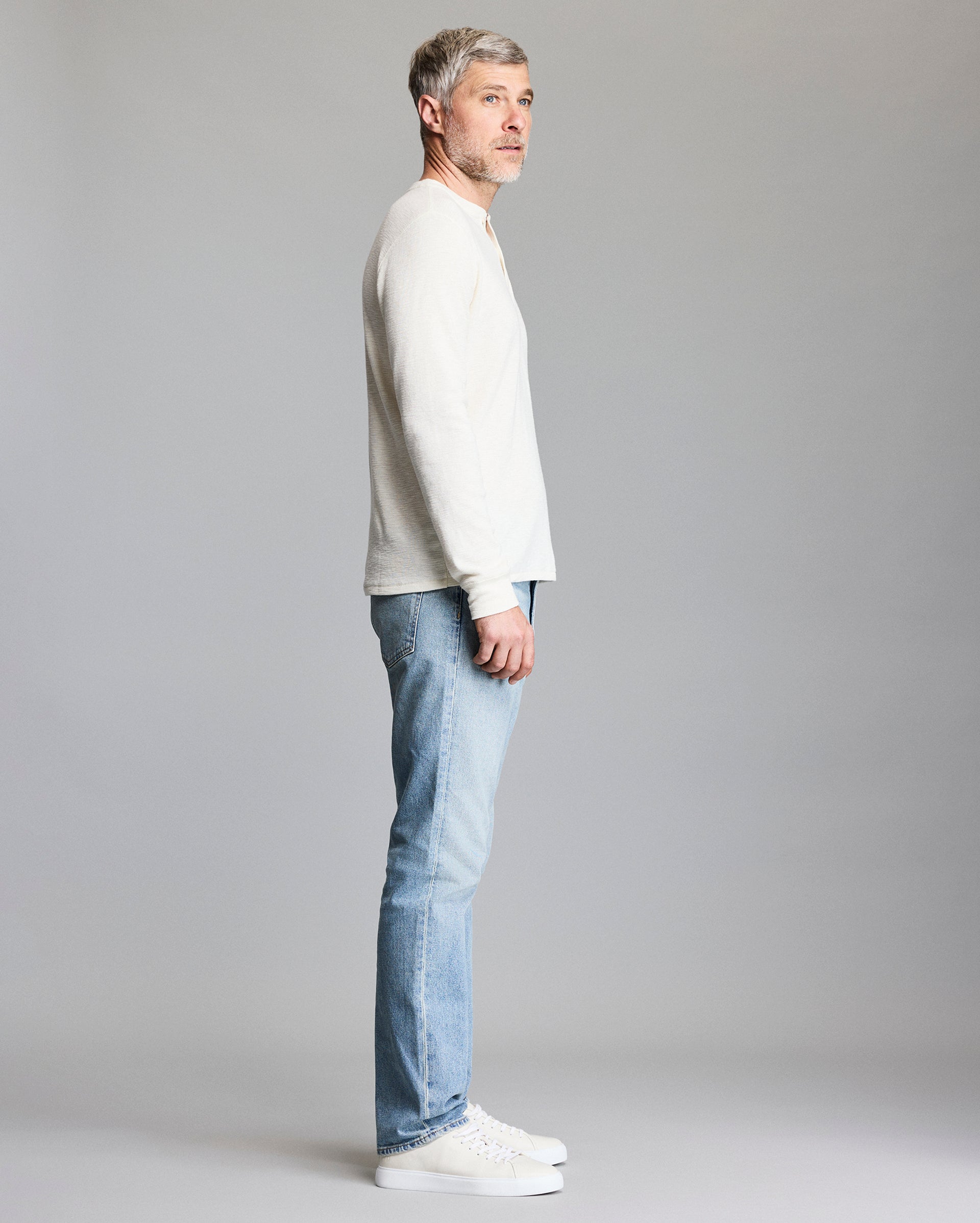 A man with short gray hair stands in profile against a plain gray background, wearing a white long-sleeve shirt, light blue jeans, and white sneakers.