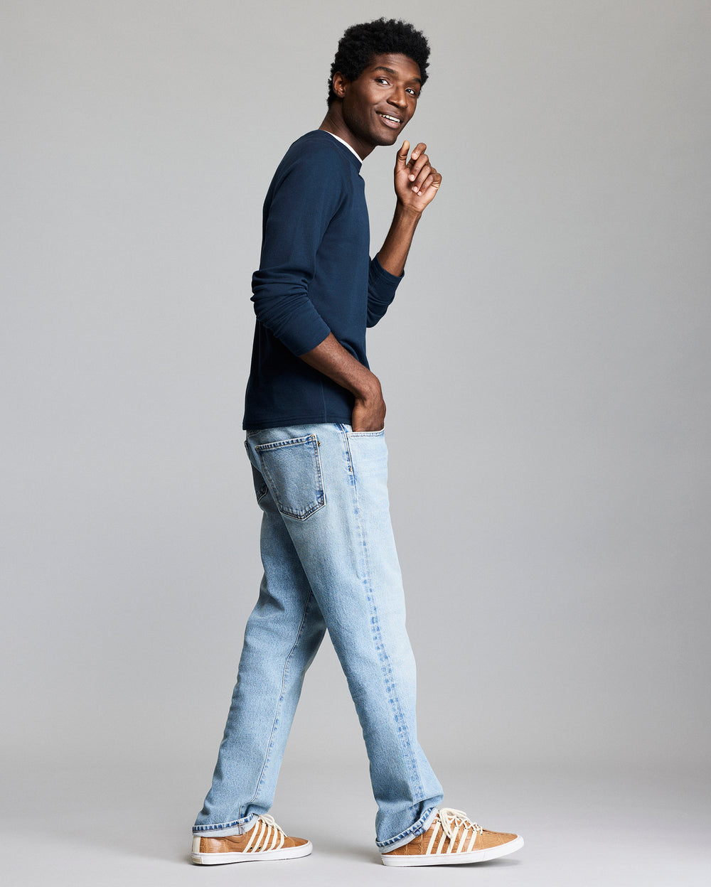 A man stands sideways, smiling and looking over his shoulder. He wears a navy long-sleeve shirt, light blue jeans, and brown sneakers with white stripes, against a plain light gray background.