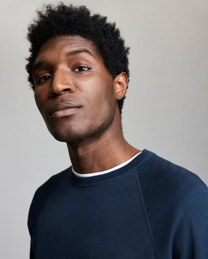 A young man with short, curly hair wearing a navy blue sweatshirt over a white shirt looks confidently at the camera against a plain, light gray background.