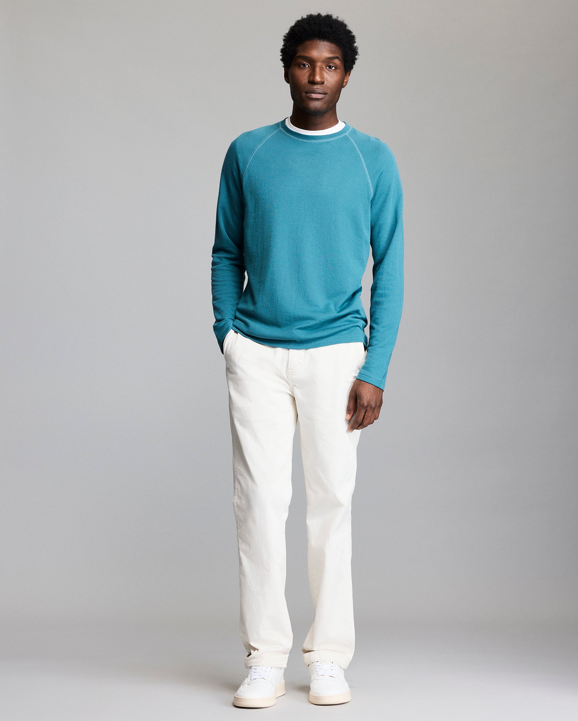 A man stands facing forward against a plain gray background, wearing a teal long-sleeve shirt, white pants, and white sneakers. His hands are in his pockets and he has a neutral expression.