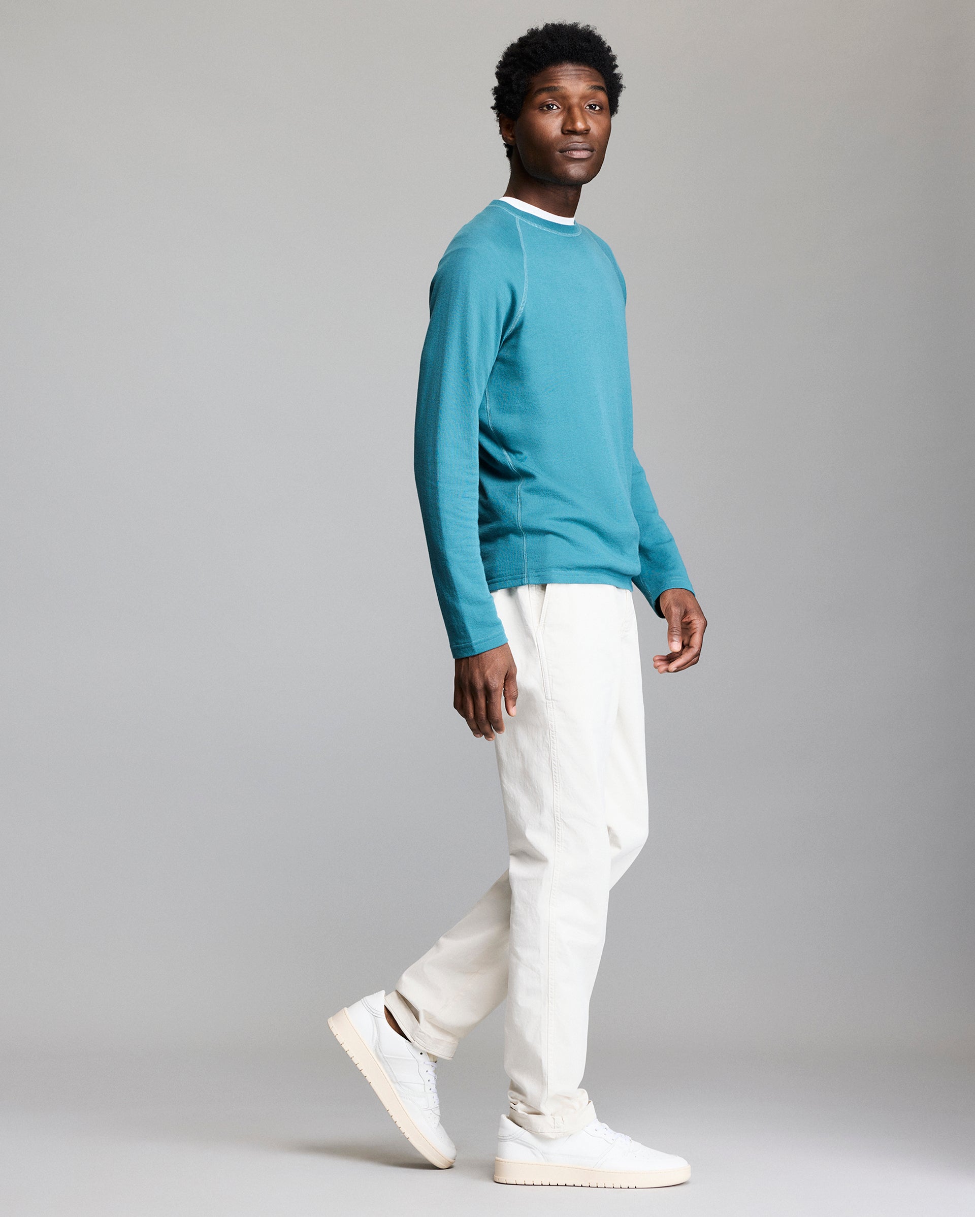 A man poses in profile against a plain background, wearing a teal long-sleeve shirt, white pants, and white sneakers. He is mid-step, with one foot slightly raised, and looking toward the camera.