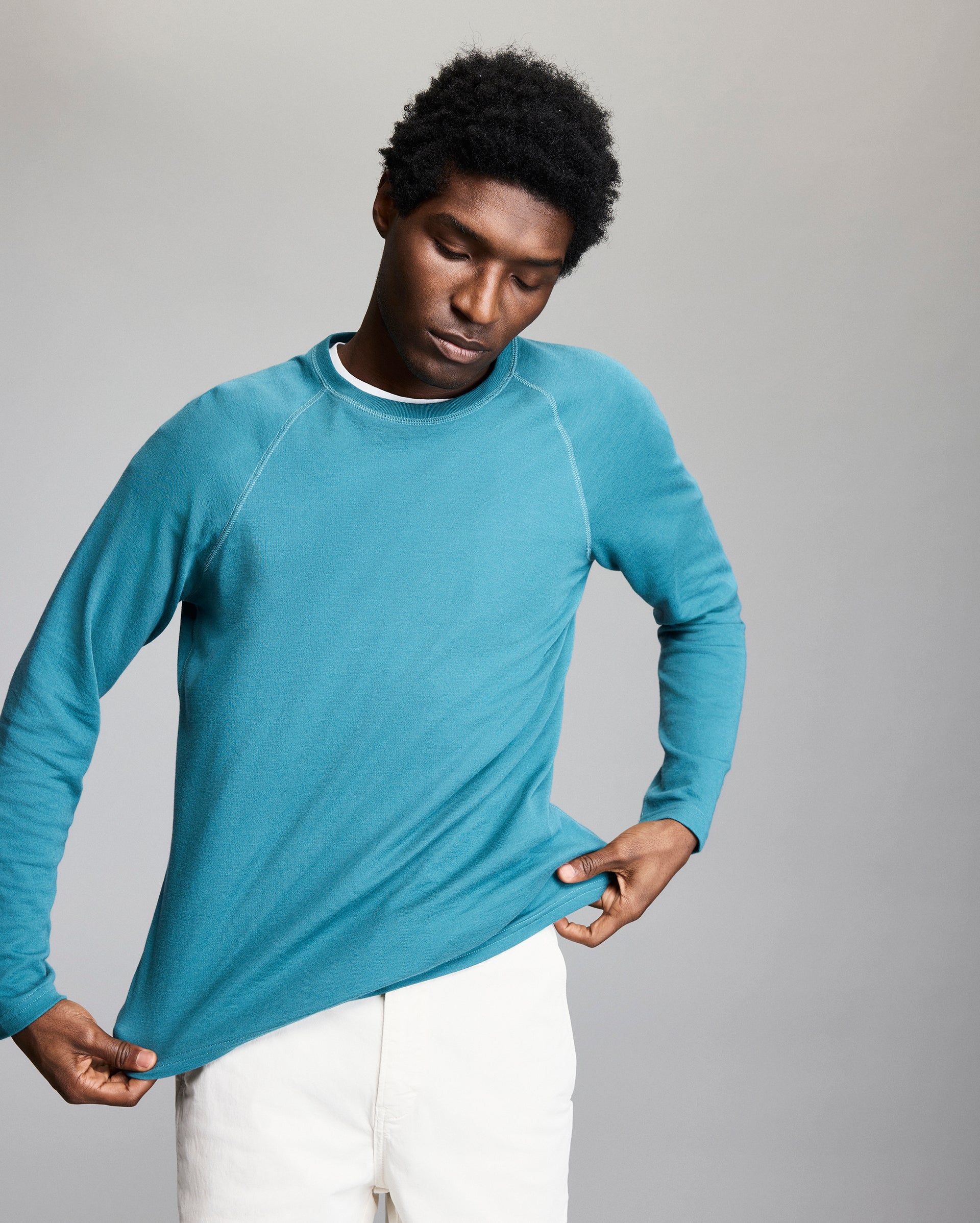 A person wearing a teal long-sleeve shirt and white pants looks down while gently pulling on the hem of the shirt against a plain gray background.