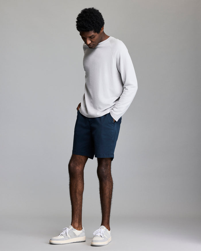 A man stands against a plain background, looking down with his hands in the pockets of his dark blue shorts. He wears a light gray long-sleeve shirt and white sneakers.