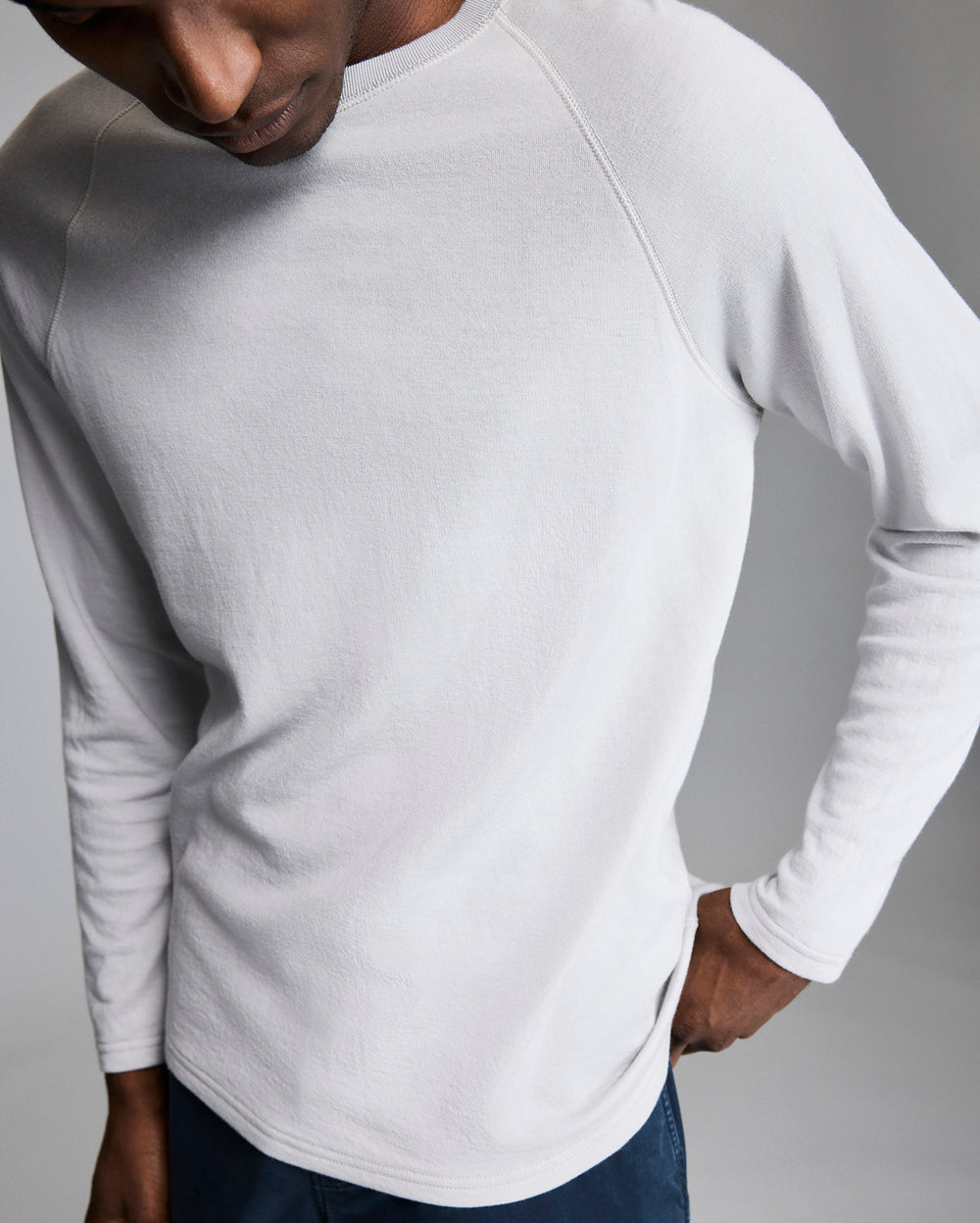 A person wearing a light gray, long-sleeve shirt and dark pants stands against a plain, neutral background, with their head turned downward and one hand resting on their hip.