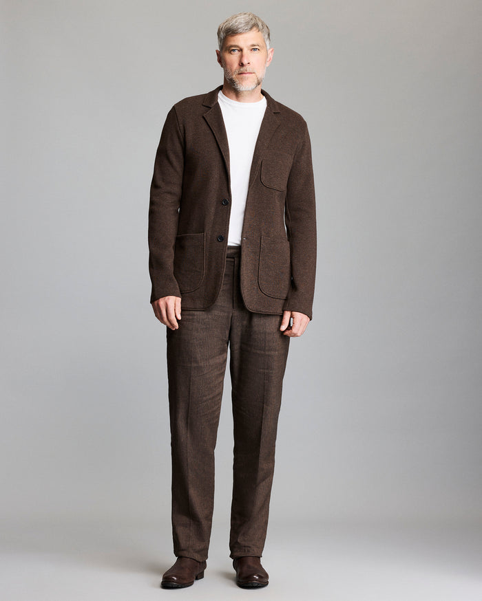 A man with short gray hair and beard wears a brown suit with a white t-shirt and brown shoes, standing against a plain light gray background.