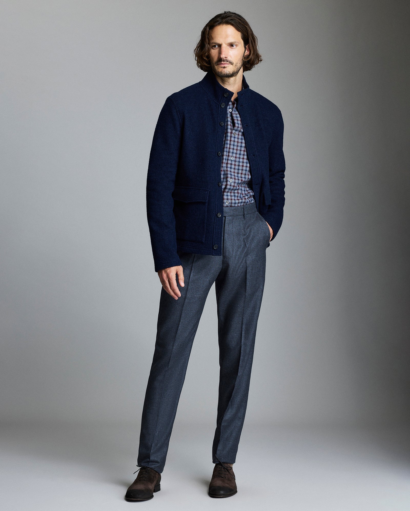A man stands in a studio wearing a dark blue jacket over a plaid shirt, gray dress pants, and brown shoes. He has medium-length hair and a neutral expression, with one hand in his pocket.
