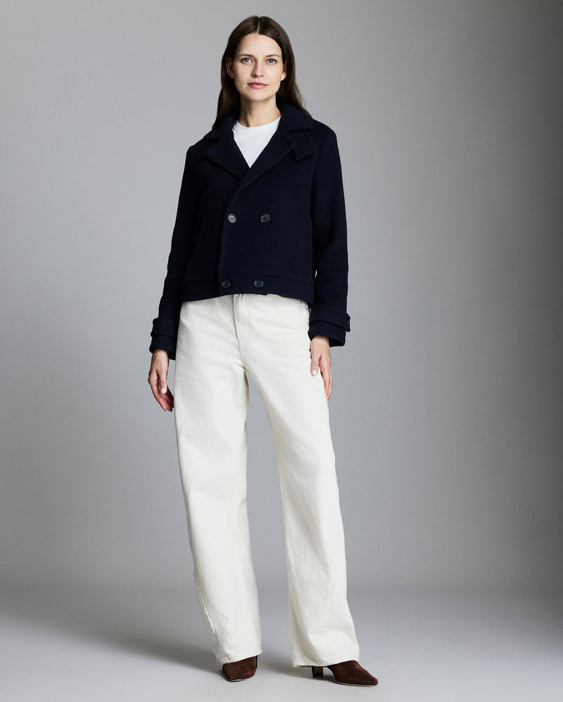 Woman wearing a dark coat and white pants against a gray background Alternate