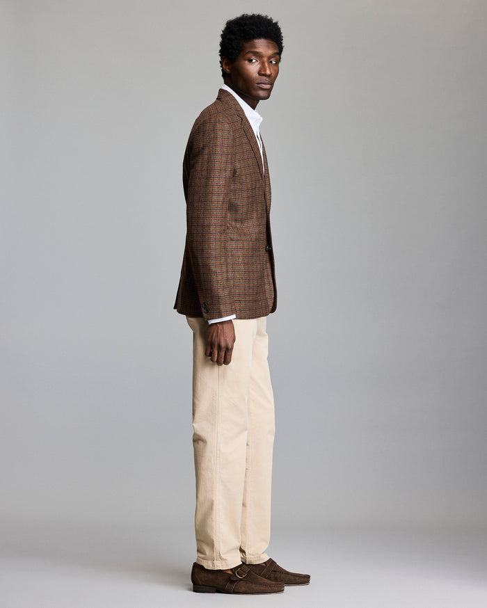 A man stands in profile against a plain background, wearing a brown plaid blazer, white shirt, beige trousers, and brown suede shoes.