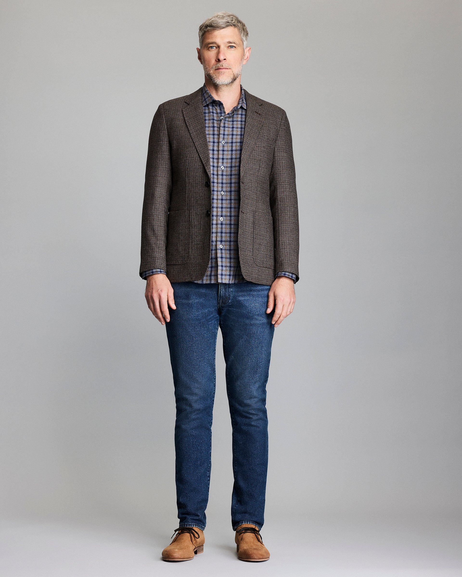Man wearing a brown checkered blazer, plaid shirt, blue jeans, and brown shoes on a gray background