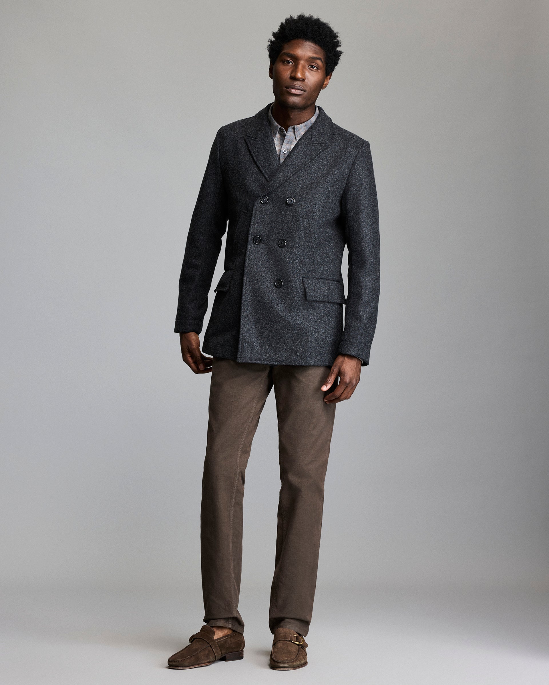 A man stands against a plain background wearing a dark double-breasted blazer, a light plaid shirt, brown pants, and brown suede loafers. He looks at the camera with a neutral expression.