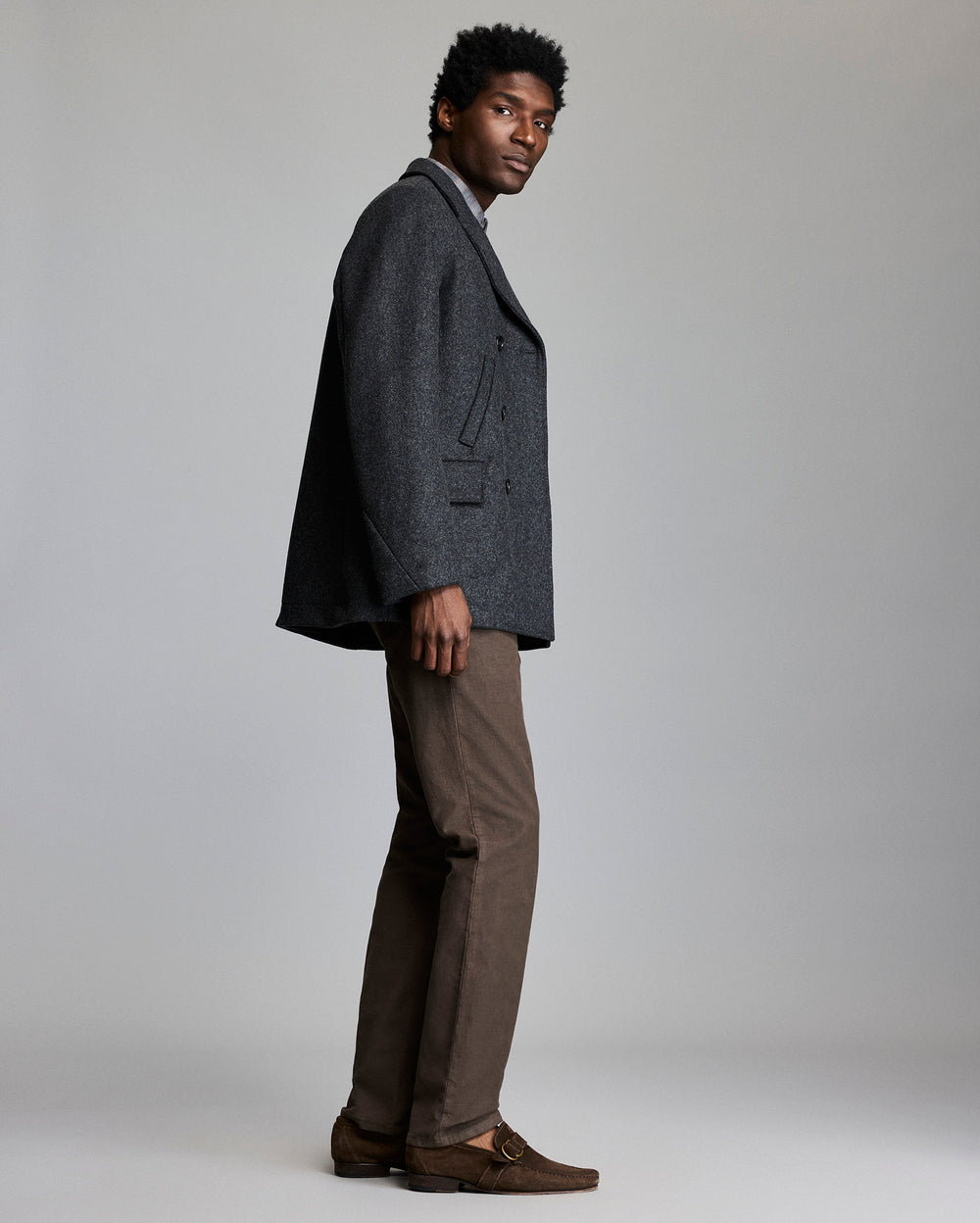 A man stands sideways against a light gray background, wearing a dark gray blazer, light gray shirt, brown pants, and brown loafers. He looks toward the camera with a neutral expression.