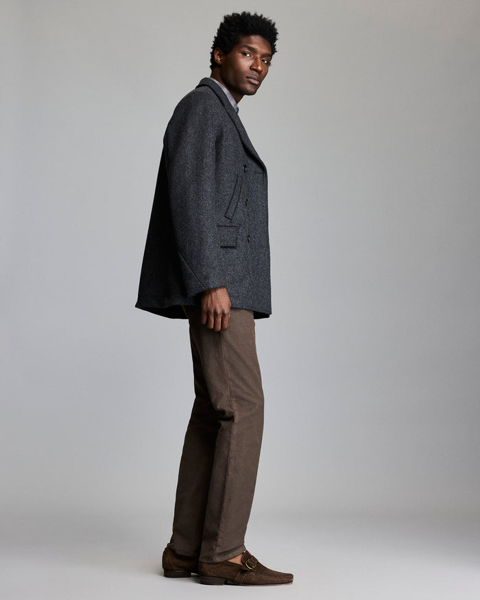 A man stands sideways against a light gray background, wearing a dark gray blazer, light gray shirt, brown pants, and brown loafers. He looks toward the camera with a neutral expression.