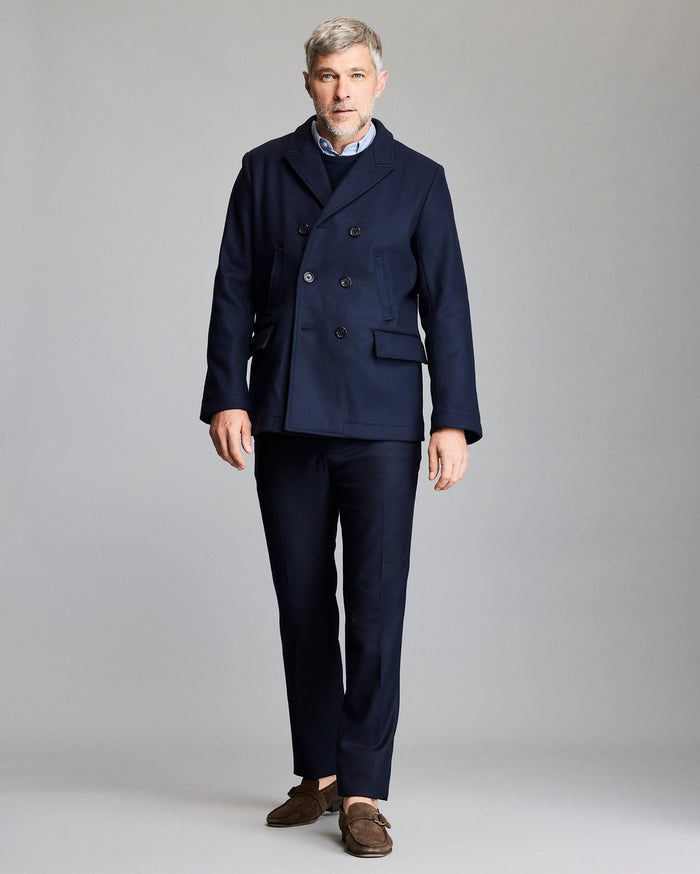 Man wearing a navy peacoat and dark pants on a gray background